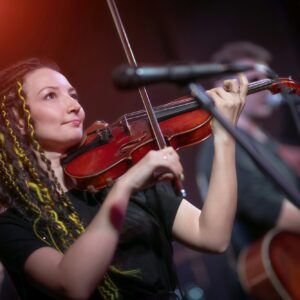 A female violinist plays the violin. Band performs on stage, rock music concert.